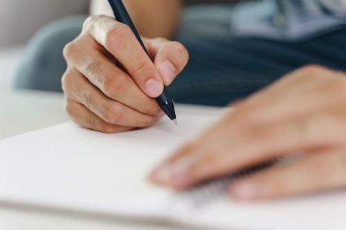 Close up of young man in casual cloth hands writing down on the notepad, notebook using ballpoint pen on the table.